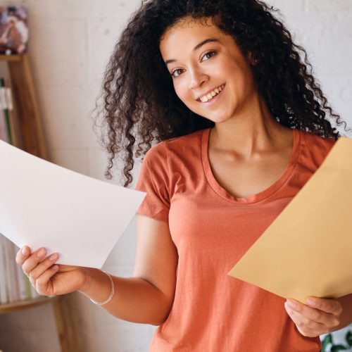 Young cheerful woman in T-shirt joyfully looking in camera holding letter with exam results in hands
